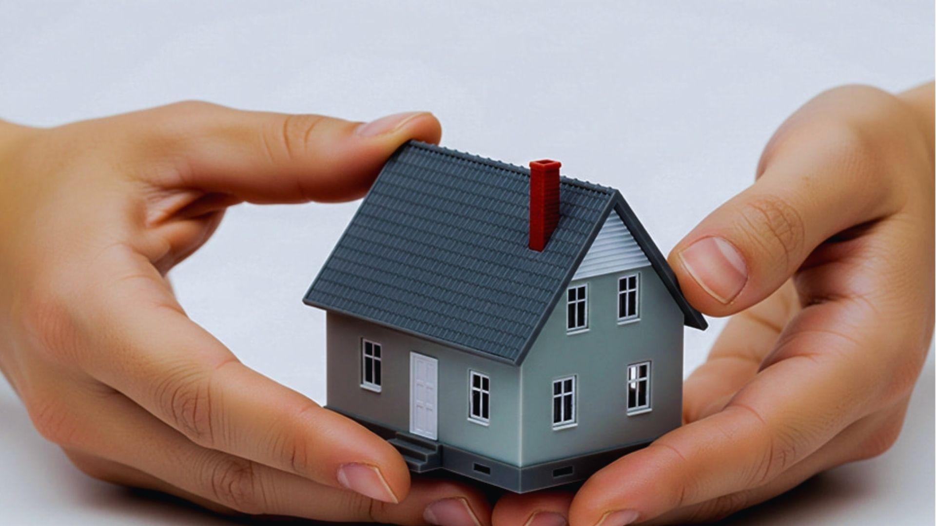 Two hands gently holding and protecting a small gray model house with a dark shingled roof and red chimney
