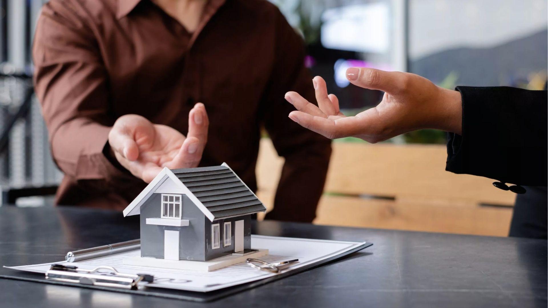 Two people gesturing over a model house and contract during a property discussion