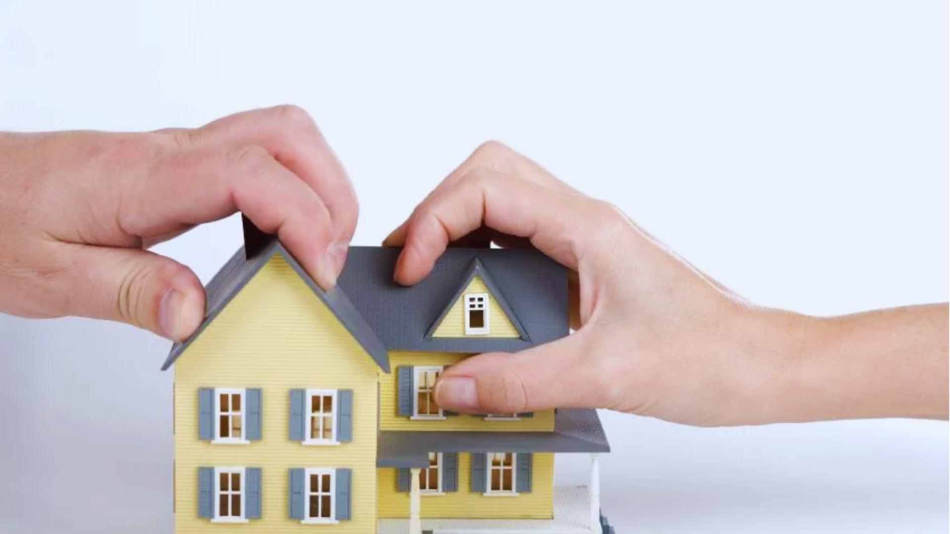 Two hands gripping a small yellow model house with gray roof against white background.