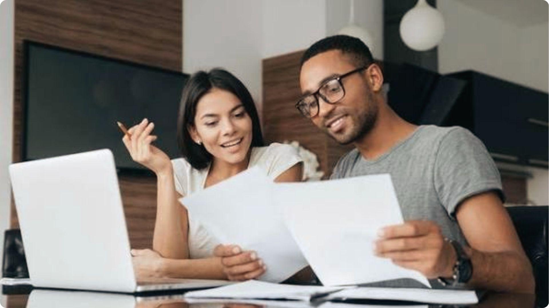 Smiling couple reviewing financial documents together at desk with laptop discussing home buying plans.