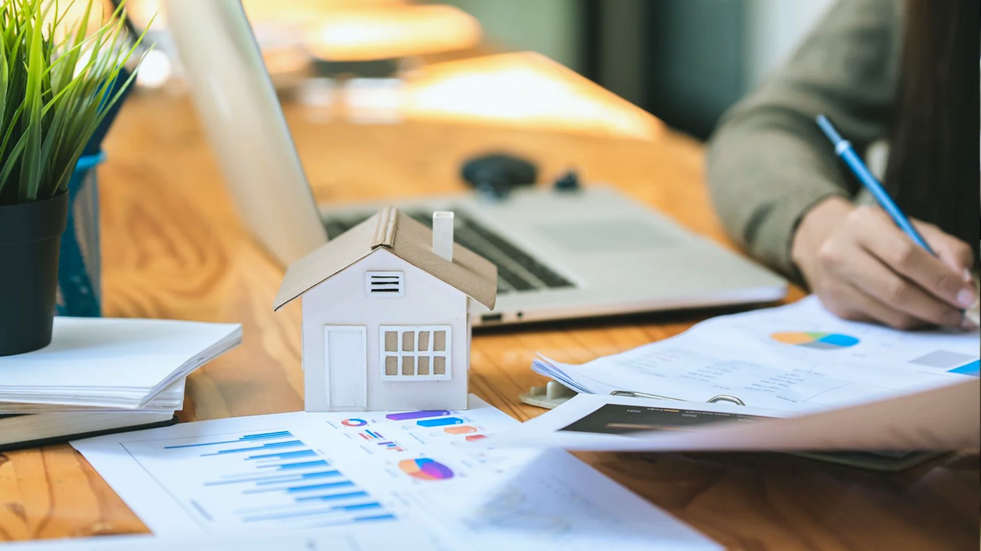 Small white house model on financial charts with person writing on property documents, real estate investment