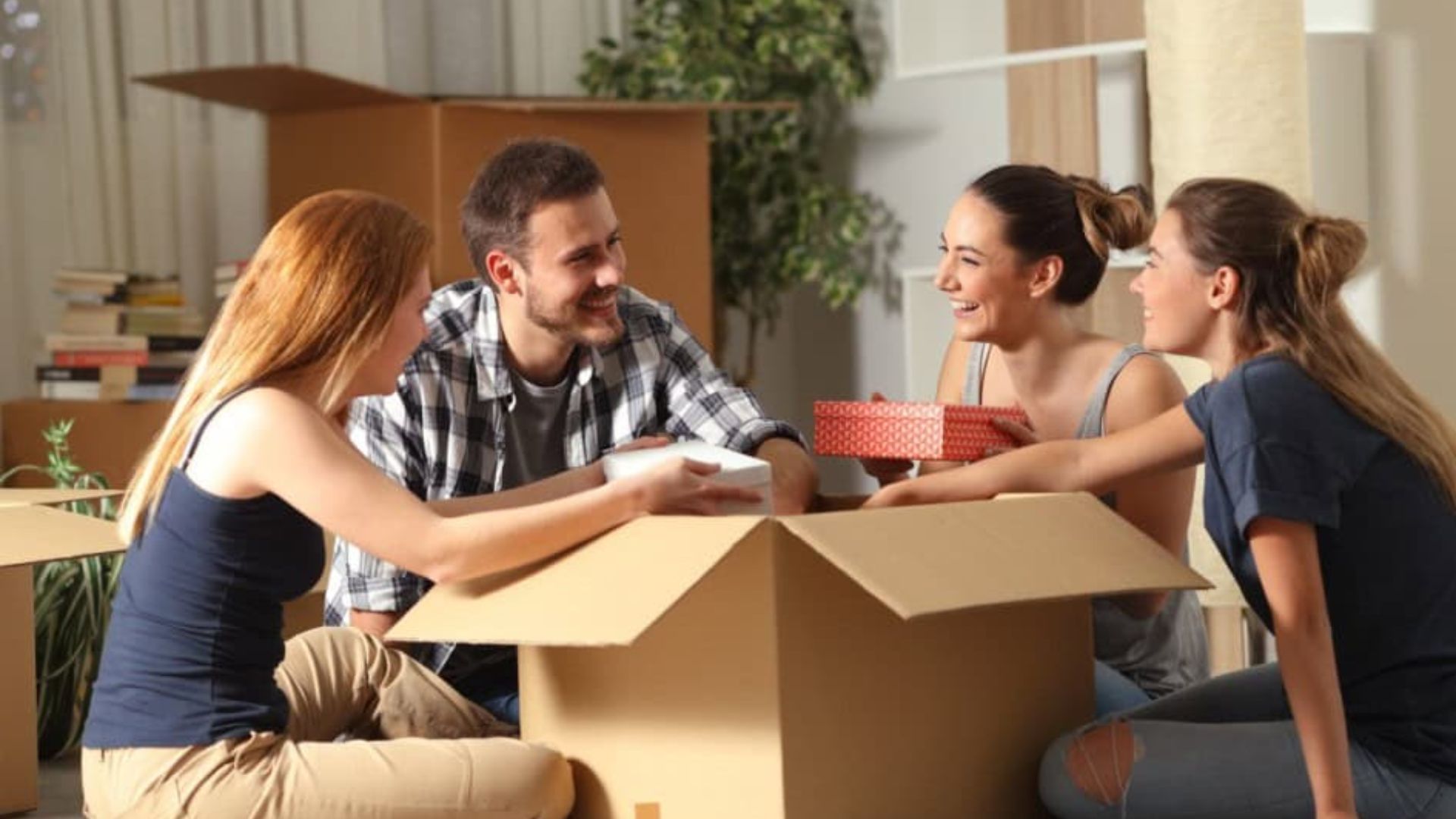 Four friends smiling while packing cardboard moving boxes with gifts on the floor in the living room, cheerful moving day.