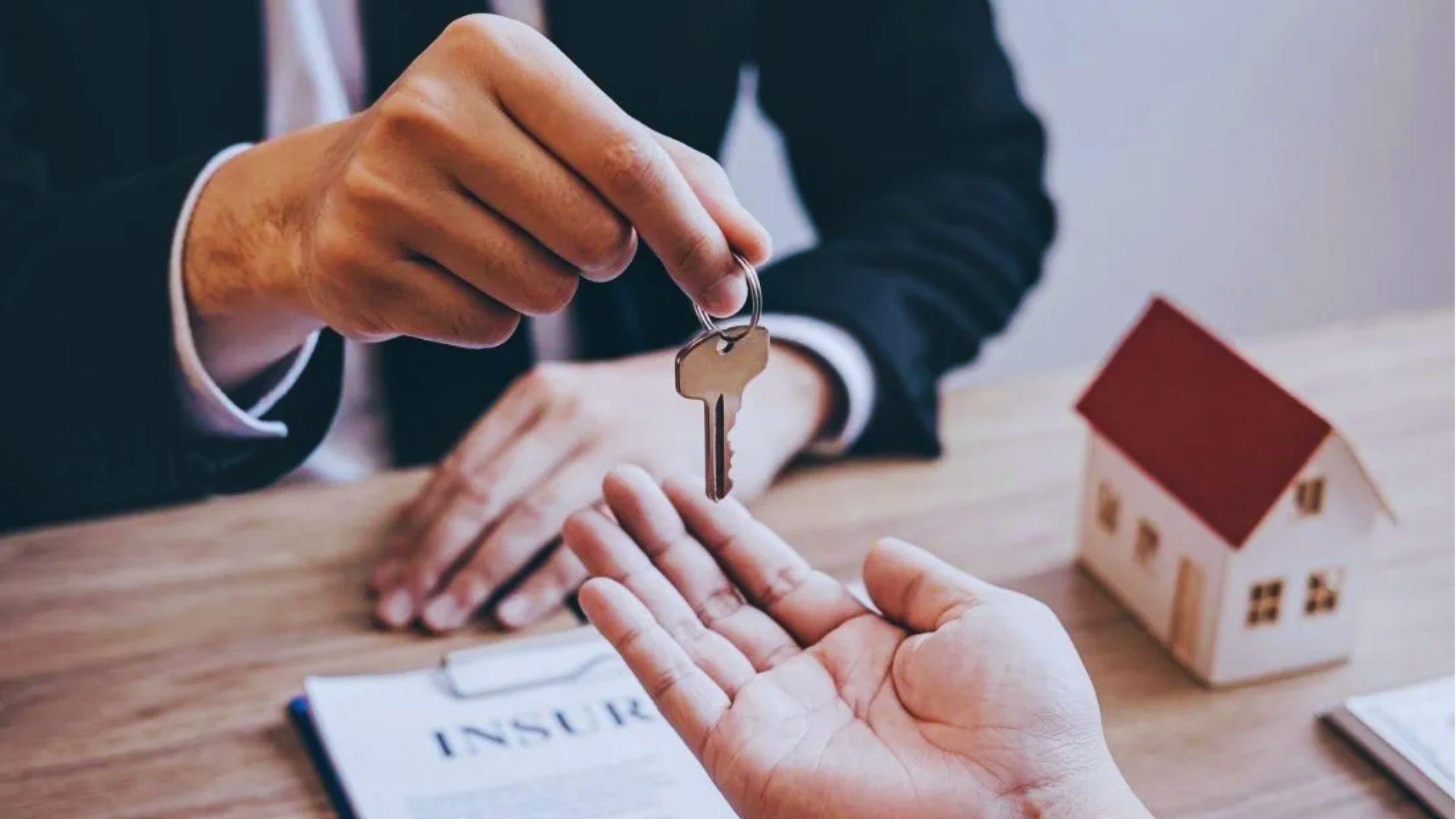 Person handing a house key to an outstretched hand over insurance documents, small model house on desk.
