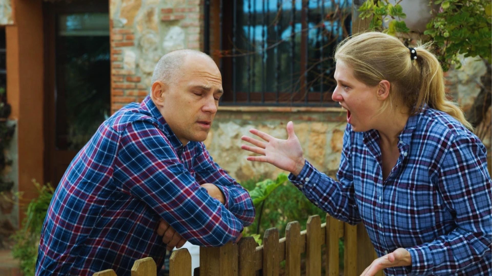 Angry neighbors arguing loudly over wooden picket fence during heated property line boundary dispute outside.