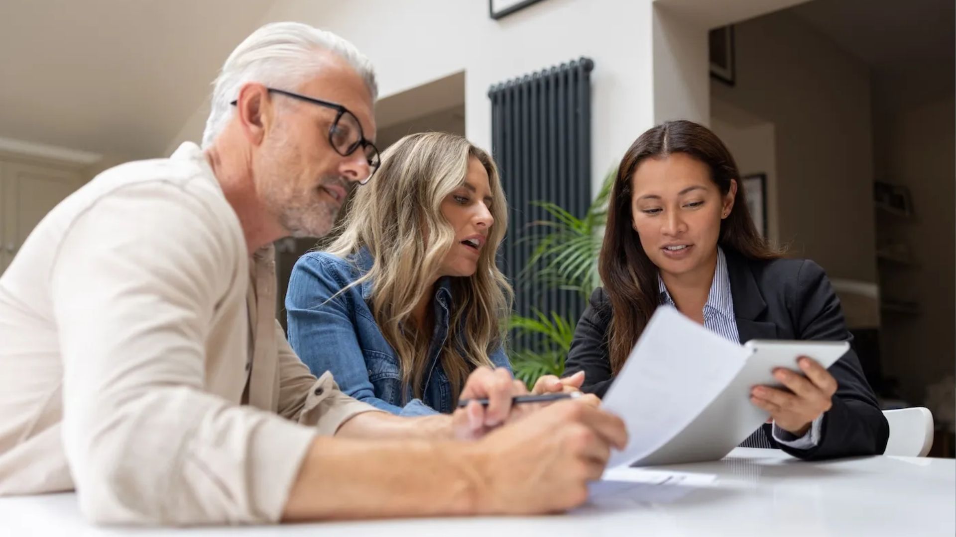 Older couple reviewing papers with a female real estate agent holding a tablet and document at a white table indoors.