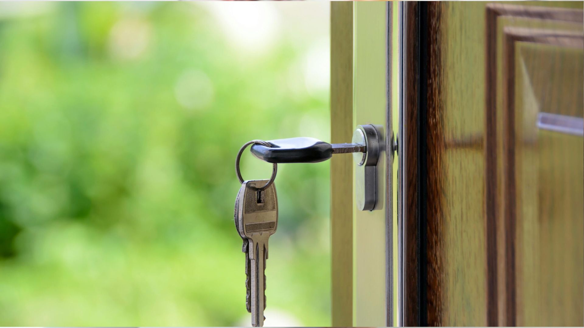 Set of silver house keys hanging from lock inserted in wooden door with blurred green garden background.
