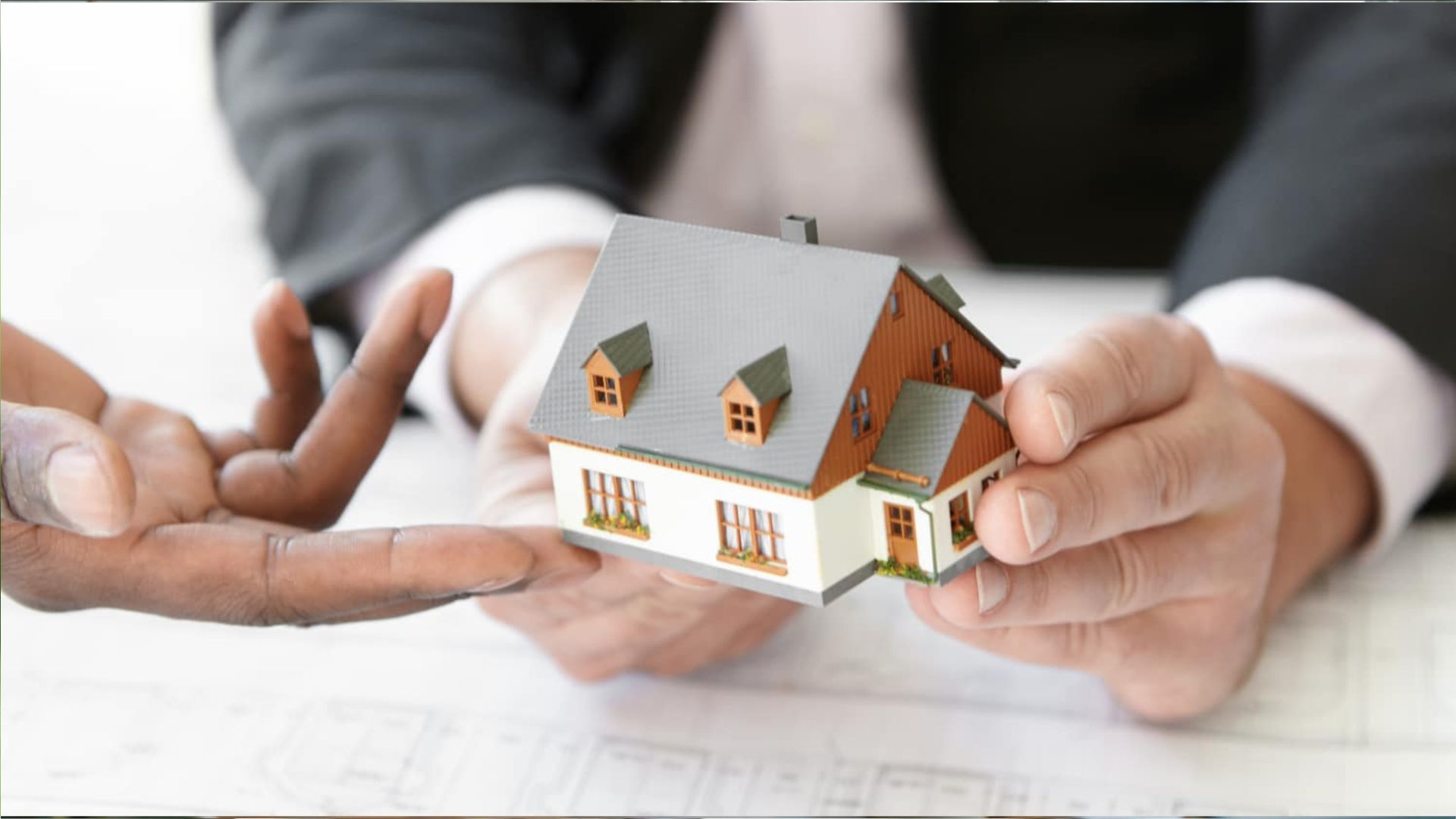 Close-up of two people exchanging small model house with gray roof and brown siding over blueprint documents.