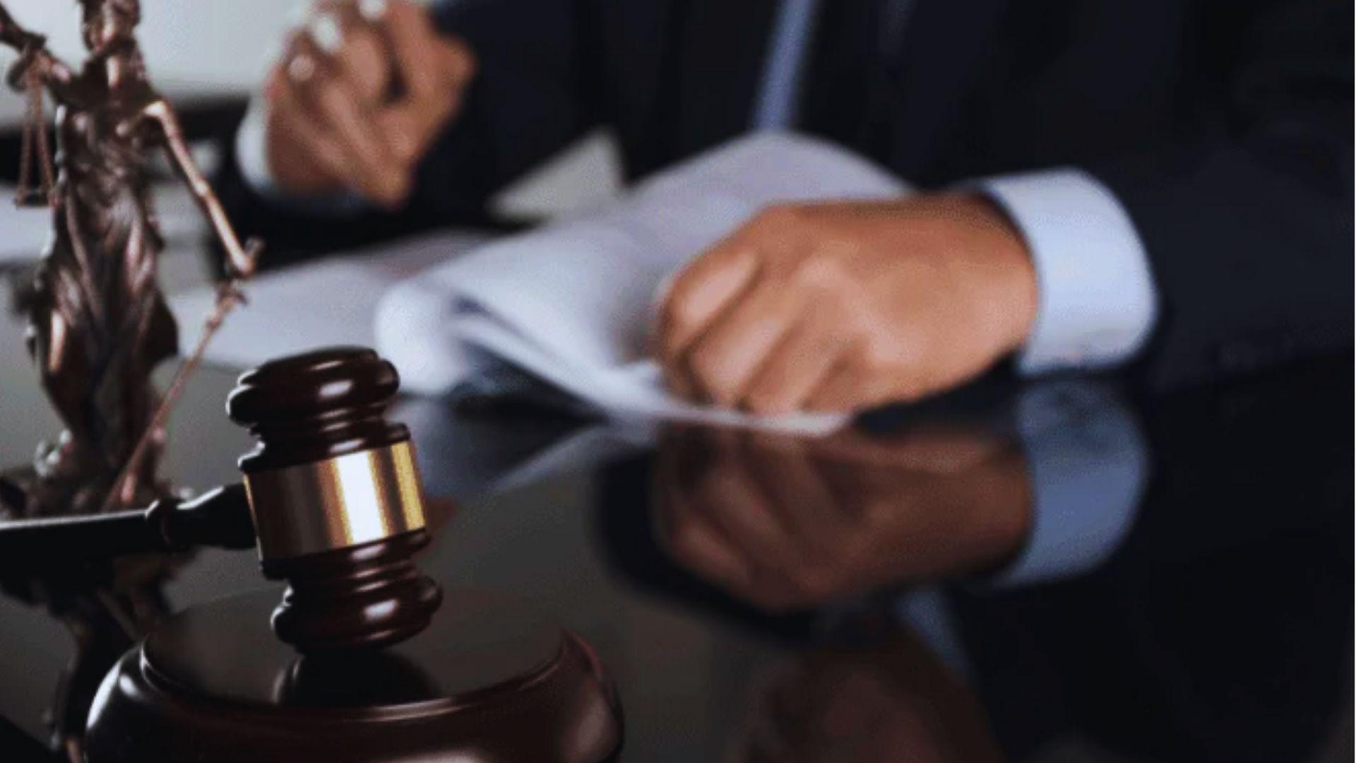 Gavel and Lady Justice statue on reflective desk, person in suit holding papers in blurred background.