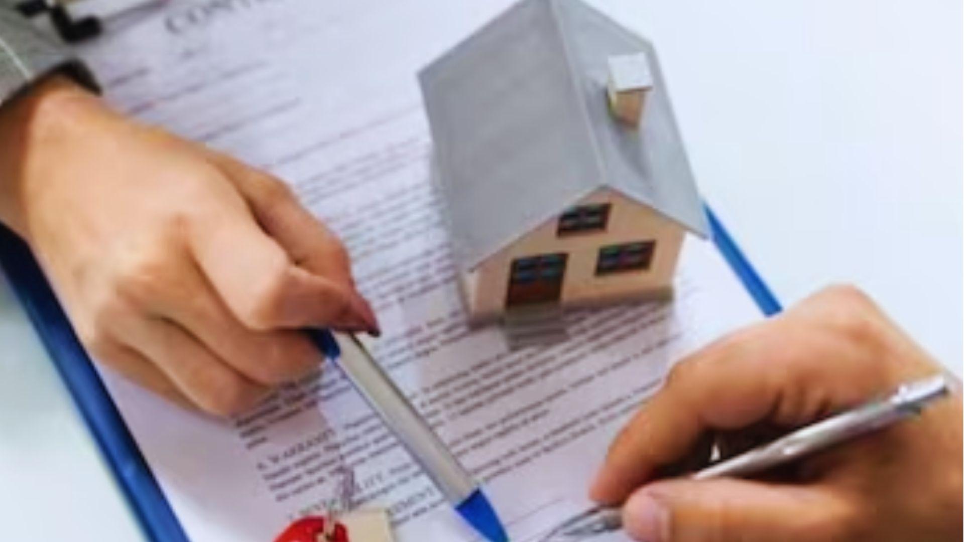 Two hands signing a contract on a clipboard with a small model house and keys on the paperwork.