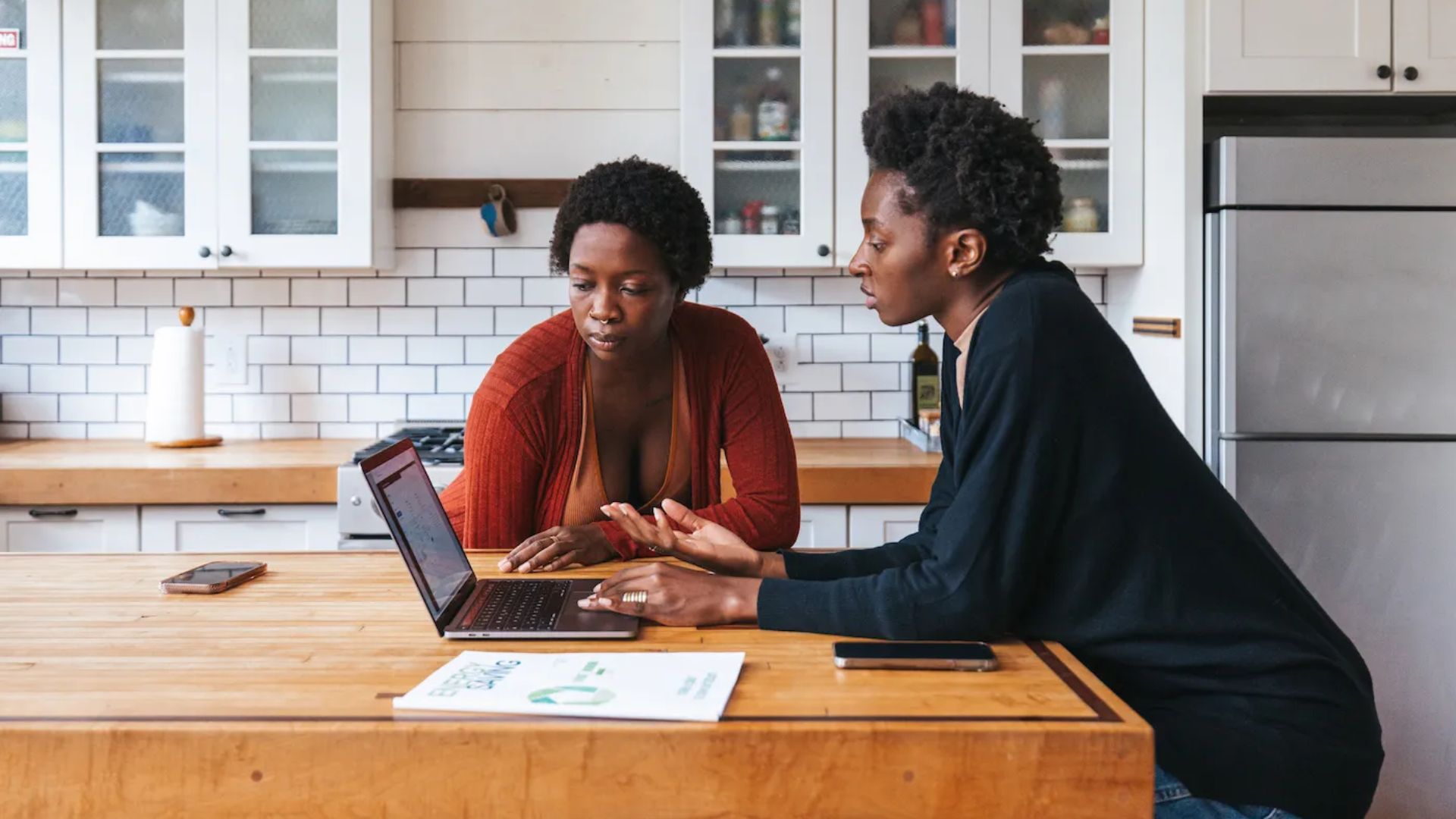 Two women leaning over kitchen island looking at laptop together with paper, phones and document titled BUSINESS on counter.