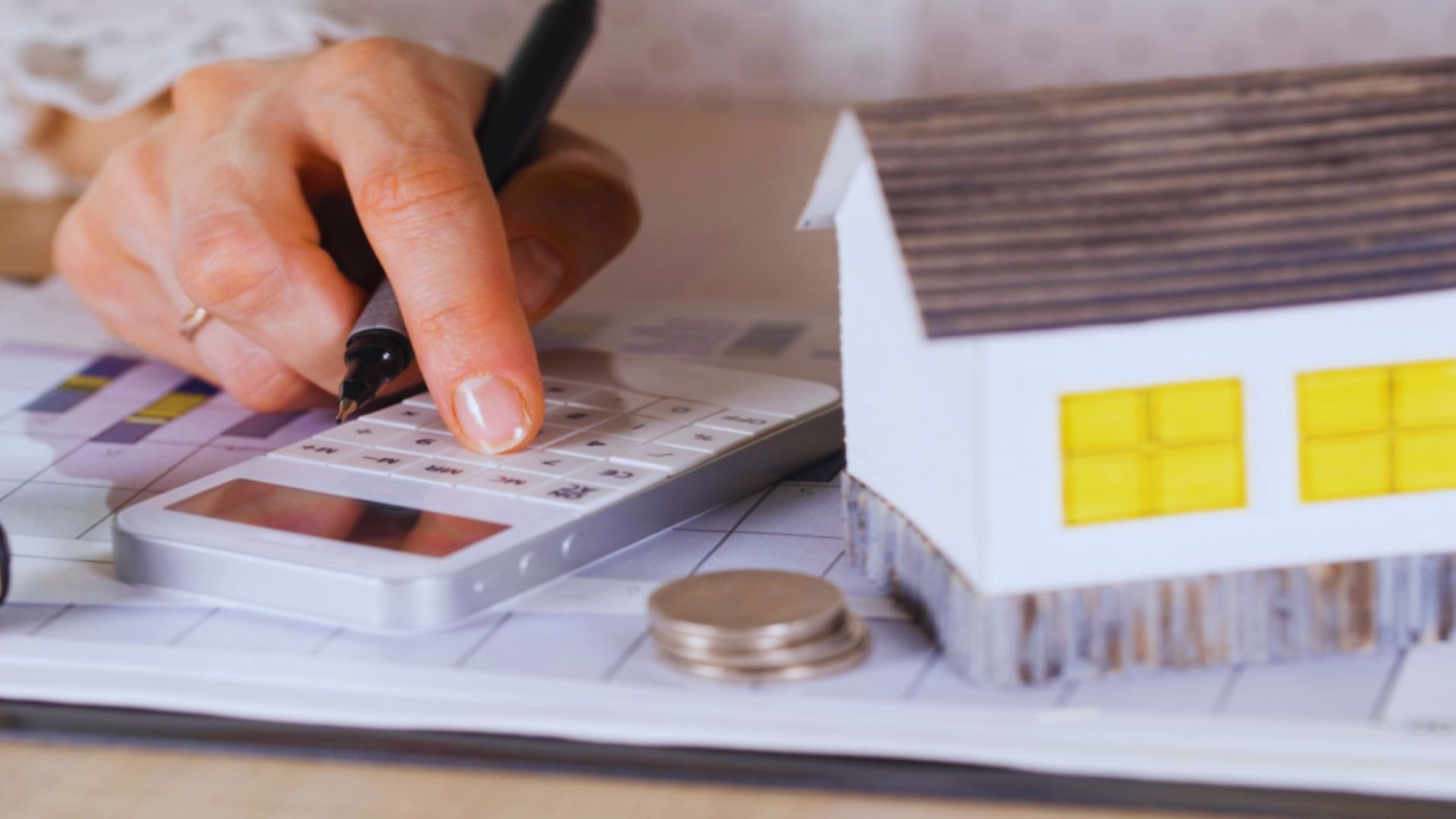 Hands using a calculator over financial papers next to a miniature house model and coins.