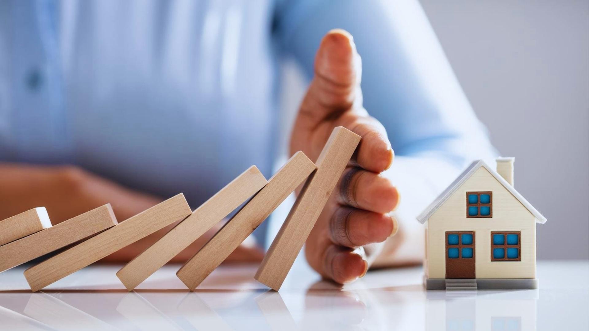 Hand blocking falling wooden dominoes from hitting small model houses protecting against foreclosure risk today.