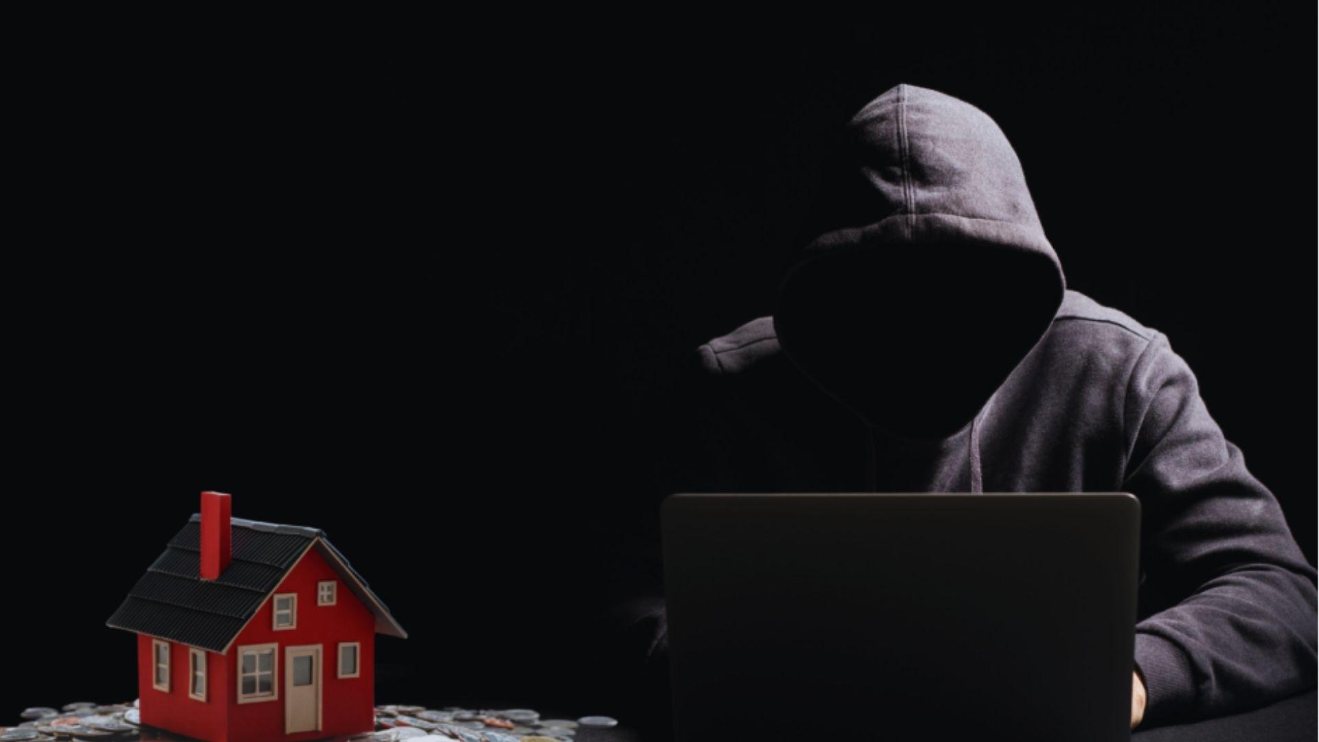 Hooded person at laptop next to small red model house on scattered coins, black background.