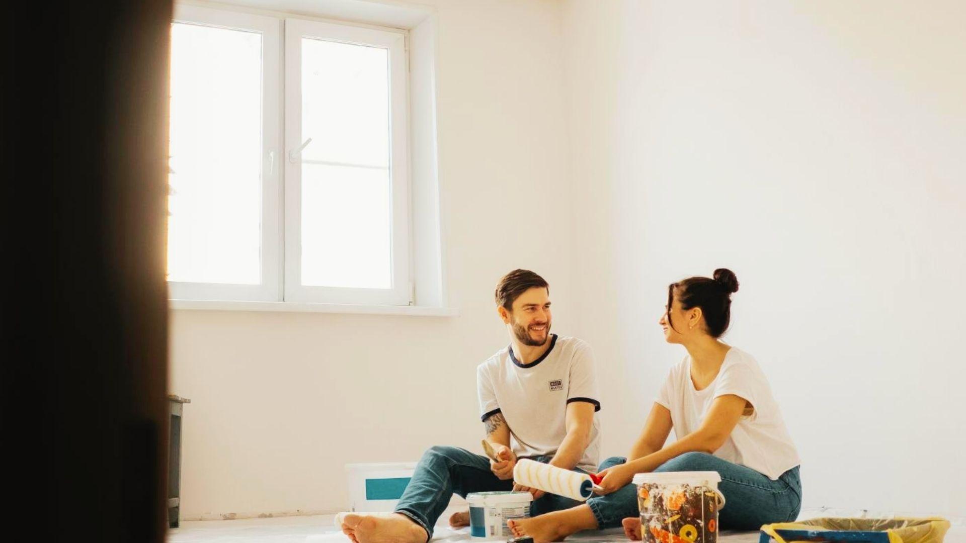 A happy couple sitting on the floor with a paint roller and buckets, smiling at each other in a bright empty room.