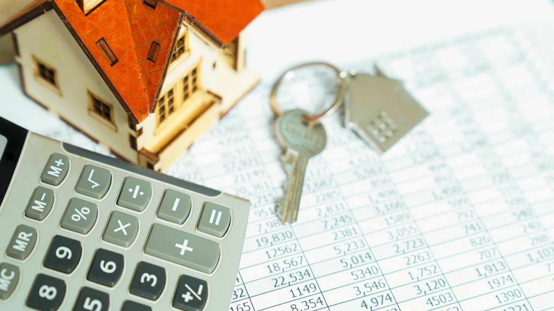 Wooden model house, house keys, and calculator placed on financial spreadsheet with numbers.