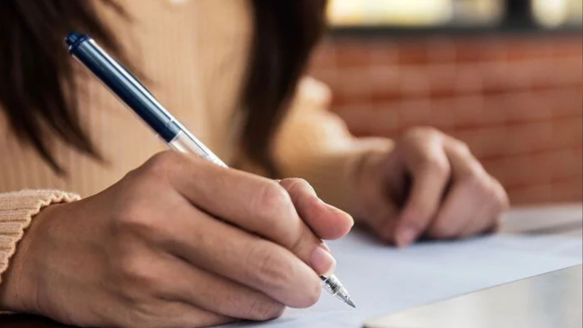 Close-up of woman's hand writing with dark blue pen on paper, beige sweater sleeve, blurred brick background.