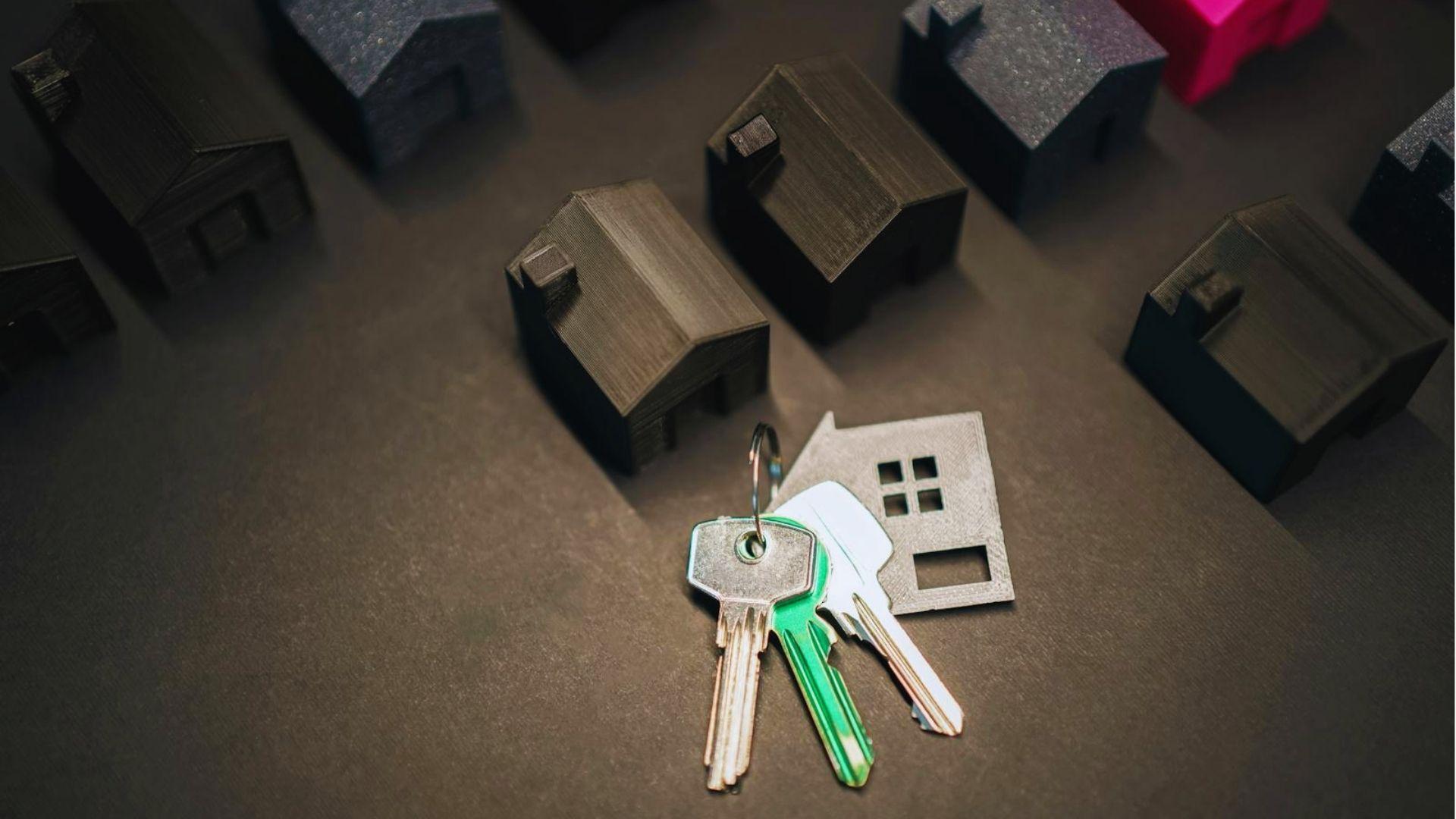 Top-down view of small black 3D-printed model houses on a dark surface, with a silver flat house cutout and a set of house keys.
