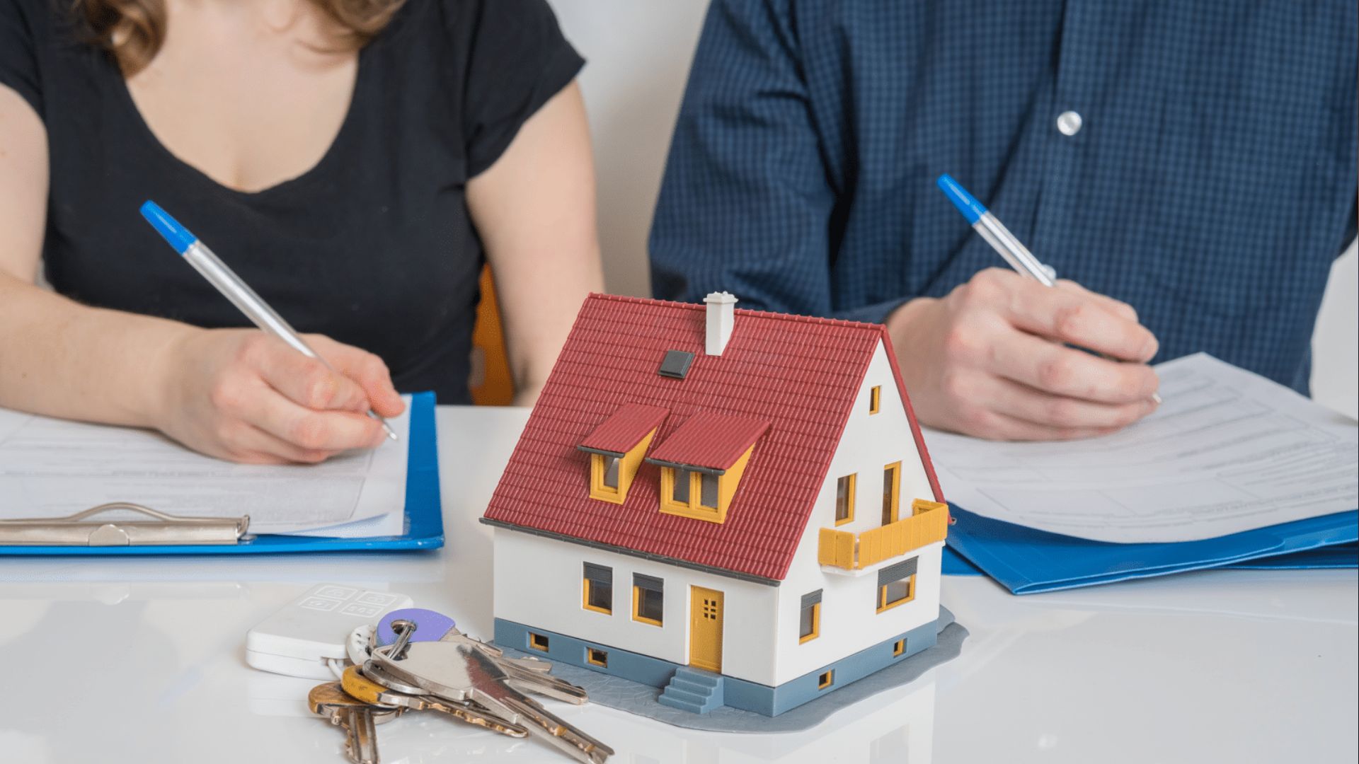 Couple signing documents with blue pens around small house with red roof and yellow trim, keys on table foreground.