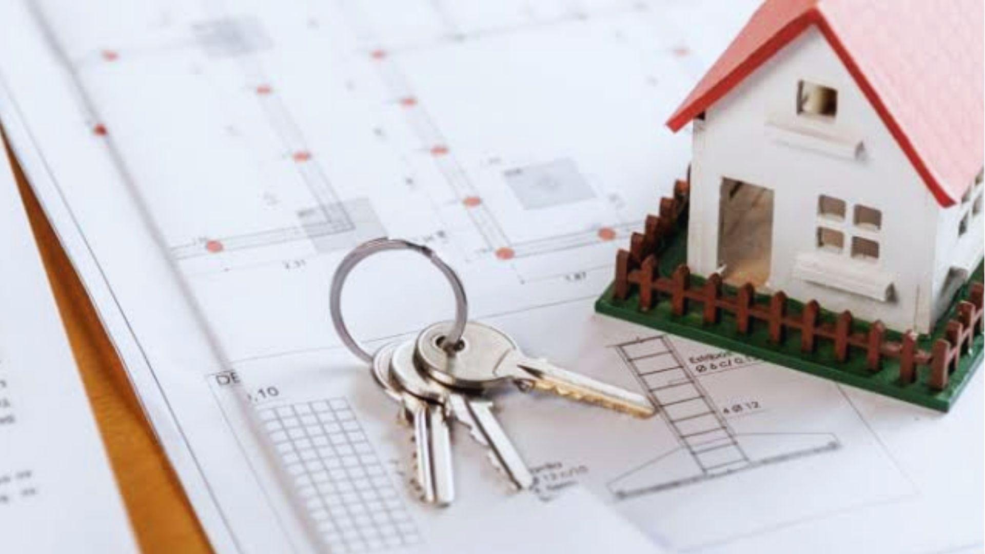 White model house with red roof and tiny brown fence sitting on architectural blueprints next to a set of silver house keys.