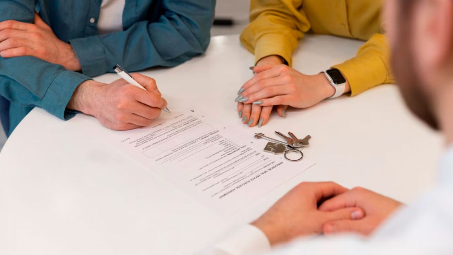 Close-up of hands signing home purchase contract on white table with set of house keys next to papers.
