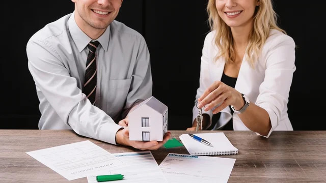 A man holds a small house model while a woman holds house keys at a desk with documents
