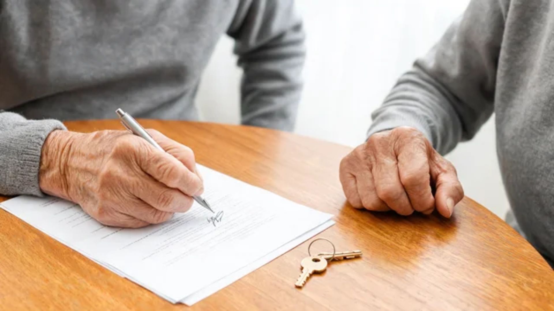 Two people at a wooden table, one signing a property deed with a pen while a house key lies beside the document